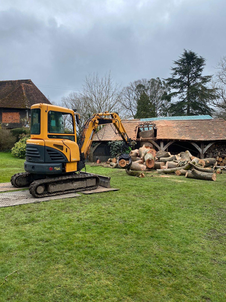 This is a photo of a country house, and the outbuilding has had a tree growing through its roof. The tree is currently being removed in the photo, and there are sections of the tree stump on the ground in front of the building. There is also a JCB which is being used to lift the sections of trunk. Photo taken by Lowestoft Tree Surgeons.